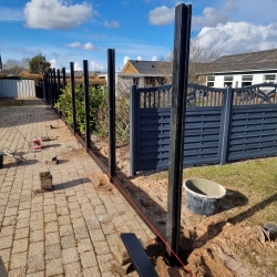 Mørke hegnstolper sat op langs en flisebelagt sti i haven. Hegnet er under opførelse i Aalborg på en solrig dag med blå himmel.