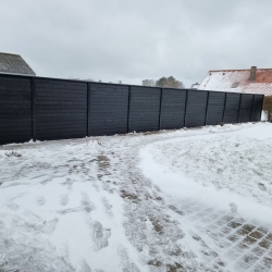 Mørkt hegn dækket af sne i en stor, åben have i Aalborg. Det solide hegn skaber en kontrast til den hvide sne og giver privatliv.