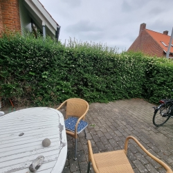 Terrasse med hæk og hegn i baggrunden, omgivet af murstensbygninger. En hyggelig udendørs plads i Hjørring med cykler og havemøbler.