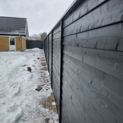Nyt sort hegn sat op i Hjørring langs flisebelægning i en åben have. Hegnet giver et moderne og minimalistisk udseende til det udendørs område.