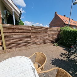 Terrasseområde med borde og stole, afgrænset af et lyst træhegn. Solrig dag og udsigt over en hyggelig have i Aalborg.