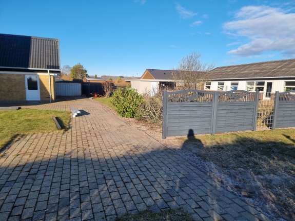 Have med delvist færdiggjort hegn i Hjørring på en solrig dag. De mørke stolper og begyndelsen af hegnet står i kontrast til den blå himmel og grønne have.