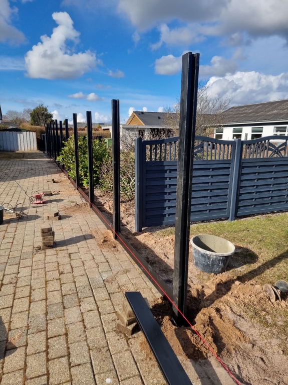 Mørke hegnstolper sat op langs en flisebelagt sti i haven. Hegnet er under opførelse i Aalborg på en solrig dag med blå himmel.