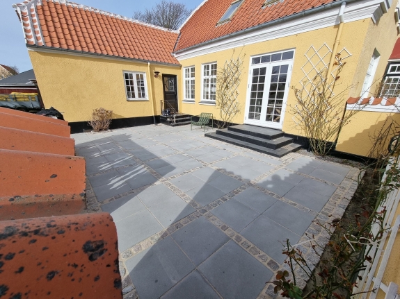 Flisebelagt terrasse ved et gult hus i Aalborg, med rene linjer og moderne belægning, der giver et pænt og funktionelt udendørsområde.
