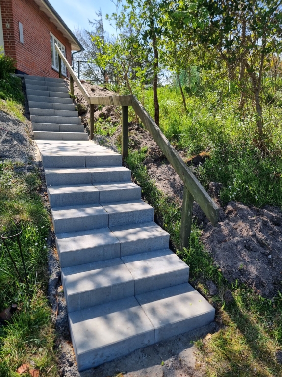 Trappe omgivet af grønne omgivelser i Hjørring, der leder op til en terrasse ved husets indgang, med bredde fliser som belægning.