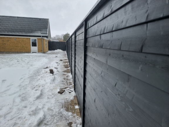 Nyt sort hegn sat op i Hjørring langs flisebelægning i en åben have. Hegnet giver et moderne og minimalistisk udseende til det udendørs område.
