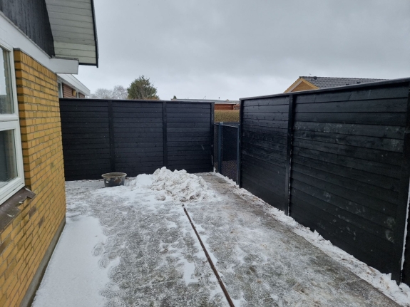 En stor have i Hjørring omgivet af et mørkt hegn, der står langs fliserne og skaber et afgrænset udendørs rum. 