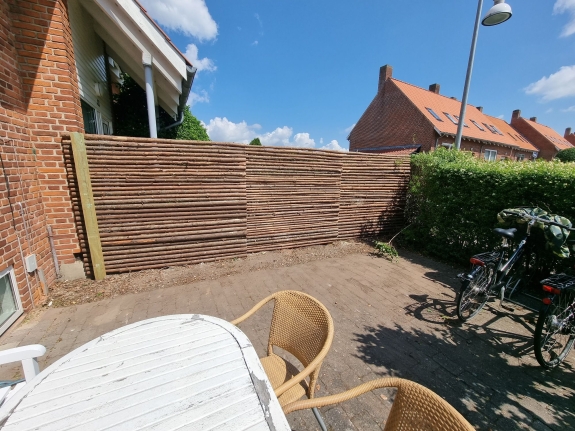 Terrasseområde med borde og stole, afgrænset af et lyst træhegn. Solrig dag og udsigt over en hyggelig have i Aalborg.